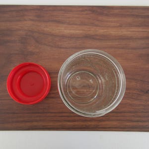 May include: An overhead shot of a clear glass jar with a red lid, placed on a dark brown wooden cutting board. The jar is empty and has a wide mouth. The lid is circular and sits to the left of the jar.