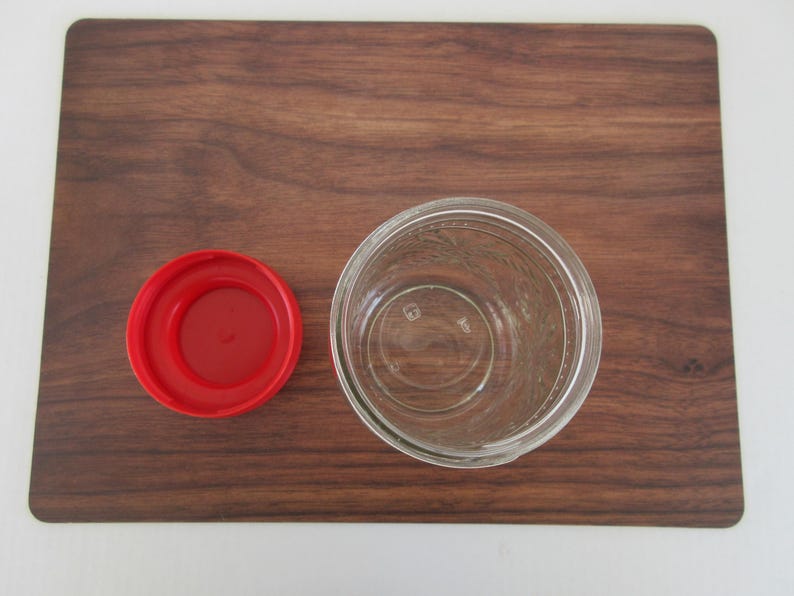 May include: An overhead shot of a clear glass jar with a red lid, placed on a dark brown wooden cutting board. The jar is empty and has a wide mouth. The lid is circular and sits to the left of the jar.
