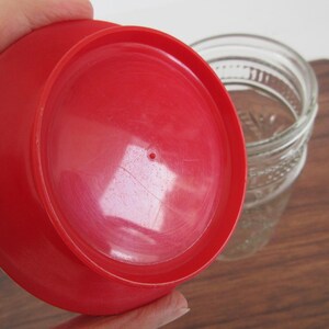 May include: A bright red plastic lid, likely for a jar, is held in front of a clear glass jar. The lid has a circular shape with a small hole in the center. The jar has a textured design.