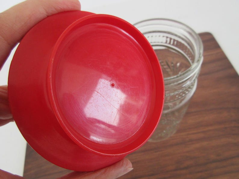 May include: A bright red plastic lid, likely for a jar, is held in front of a clear glass jar. The lid has a circular shape with a small hole in the center. The jar has a textured design.