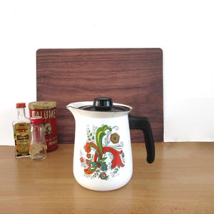 May include: White enamelware coffee pot with a black handle and lid, decorated with a colorful floral design in red, green, and orange. The pot is displayed with small vintage bottles and a wooden cutting board.