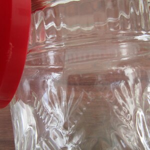 May include: Close-up of a clear glass jar with a red lid. The jar has a textured, embossed design. The lid is a solid, bright red color. The jar is partially open, revealing the interior.
