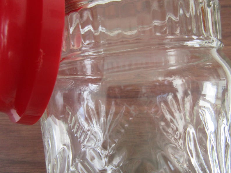 May include: Close-up of a clear glass jar with a red lid. The jar has a textured, embossed design. The lid is a solid, bright red color. The jar is partially open, revealing the interior.