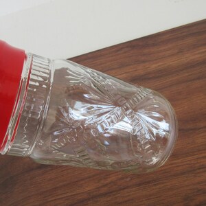 May include: A clear glass cocktail shaker with a red lid. The glass has a textured, embossed design. The shaker is angled on a dark brown wooden surface. The image is well-lit, highlighting the details of the glass.