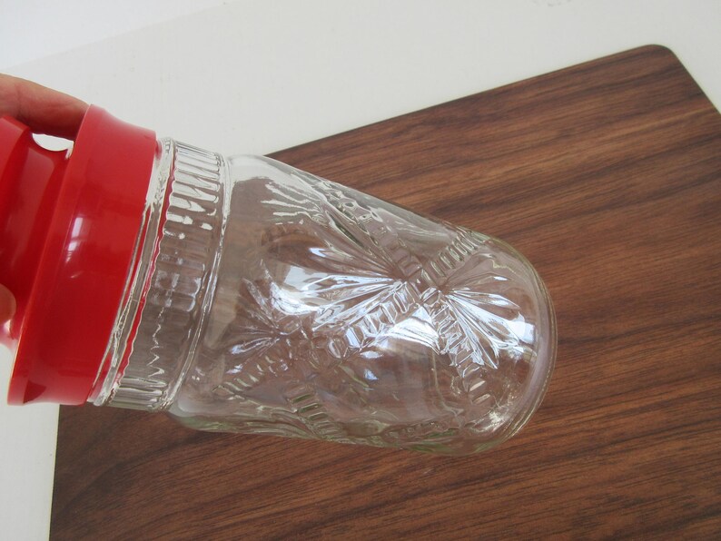 May include: A clear glass cocktail shaker with a red lid. The glass has a textured, embossed design. The shaker is angled on a dark brown wooden surface. The image is well-lit, highlighting the details of the glass.