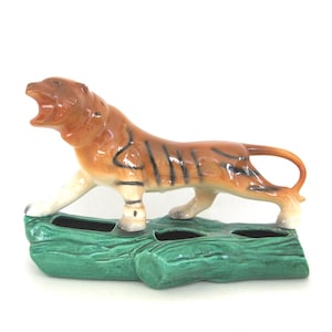 May include: A ceramic tiger figurine in shades of orange and brown with black stripes, standing on a green log-shaped base. The tiger has an open mouth and white accents on its paws.