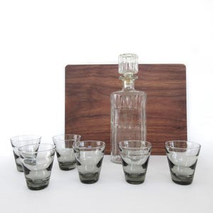 May include: A clear glass decanter with a stopper and six gray-tinted drinking glasses are arranged in front of a wooden cutting board. The glasses have a rounded shape. The decanter is rectangular with vertical lines.