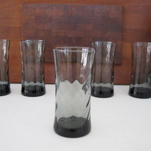 May include: Five tall, gray-tinted drinking glasses with a wavy, textured design. The glasses have a dark gray base and a clear rim. They are arranged in a row on a white surface, with a wooden cutting board in the background.