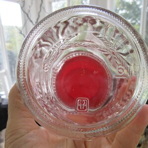 May include: Clear glass bottle with a red base. The bottle has embossed markings and a textured rim. The bottom of the bottle has a red center and a small square symbol. The bottle is held in a hand.