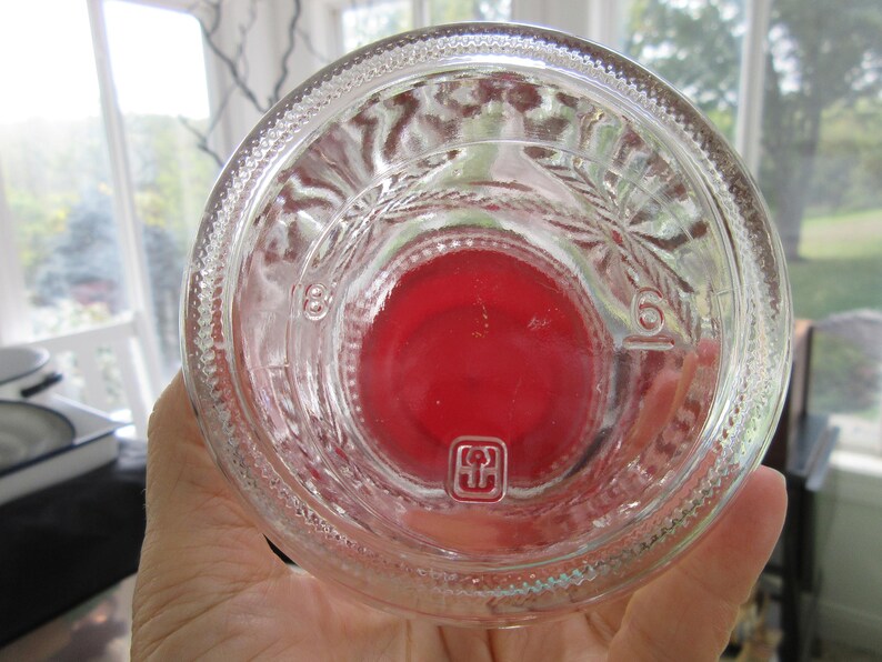 May include: Clear glass bottle with a red base. The bottle has embossed markings and a textured rim. The bottom of the bottle has a red center and a small square symbol. The bottle is held in a hand.