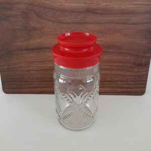 May include: A clear glass jar with a red lid. The jar has a decorative pattern and is sitting on a white surface with a wooden background. The jar is empty.