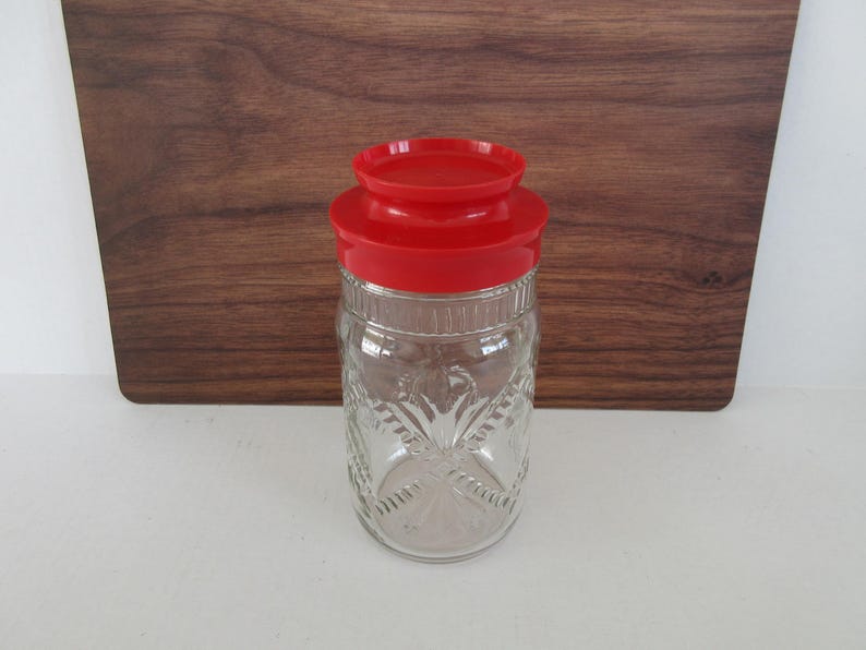 May include: A clear glass jar with a red lid. The jar has a decorative pattern and is sitting on a white surface with a wooden background. The jar is empty.