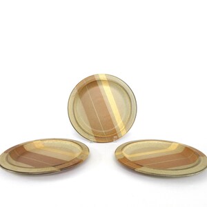May include: Three vintage ceramic plates. One is round, and two are oval. Each plate features a striped pattern in shades of brown, tan, and yellow. The plates are set against a white background.