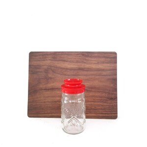 May include: A clear glass jar with a red lid, featuring a diamond pattern, stands in front of a rectangular wooden cutting board. The jar is empty and appears to be a vintage kitchen item. The background is plain white.