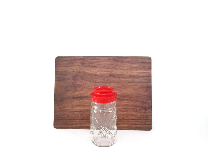 May include: A clear glass jar with a red lid, featuring a diamond pattern, stands in front of a rectangular wooden cutting board. The jar is empty and appears to be a vintage kitchen item. The background is plain white.