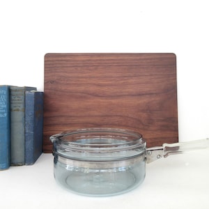 May include: A blue glass saucepan with a clear glass lid and a silver handle. The saucepan is sitting on a wooden cutting board.