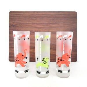 May include: Three vintage highball glasses with circus animal designs. The glasses feature orange elephants and a green lion, with a white and red decorative top and a black and white scalloped base. The background is a wooden cutting board.