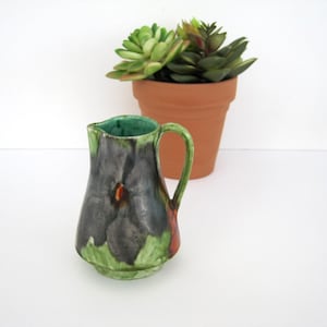 May include: A small, decorative ceramic pitcher with a dark gray and green floral design. The pitcher has a green handle and interior, with a small orange accent. A terracotta pot with green and red succulents is in the background.