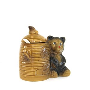 May include: A ceramic honey pot shaped like a beehive with a bear figurine on the side. The beehive is brown and has a white lid with a small spoon inside. The bear is brown and black and has a red nose.