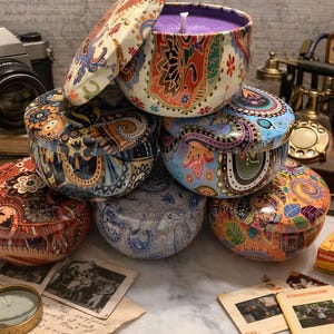 May include: A collection of decorative, patterned candle tins in various colors, including orange, blue, and purple. The tins are stacked with lids, and one is open, revealing a purple candle. Vintage photography equipment and slides are in the background.