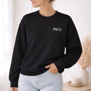 May include: Black crew neck sweatshirt with the white letters "PACU" printed on the chest. The sweatshirt is worn with light blue jeans. The image is taken indoors with neutral tones and natural light.