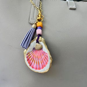 May include: A handmade seashell keychain with a gold chain and clasp. The oyster shell is painted with pink and orange stripes and has a small gold seashell charm. A lavender tassel and colorful beads complete the design.