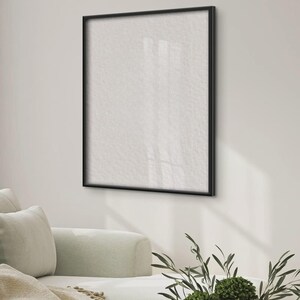May include: A square wall-mounted picture frame with a thin black border and a blank white interior surface. The frame is positioned on a plain wall above a light-colored sofa, with a small green plant visible in the foreground of the scene.