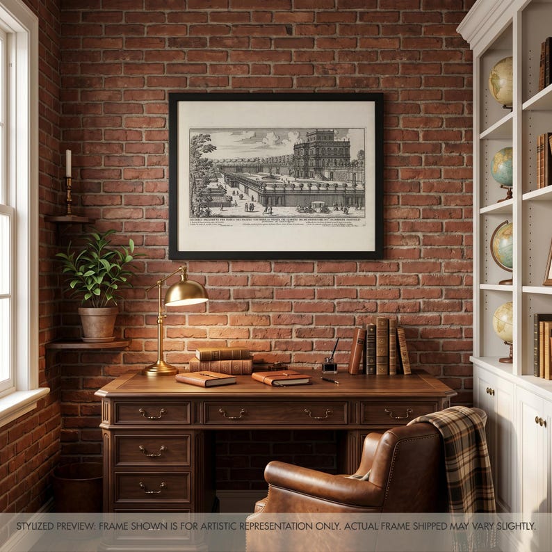May include: A black framed wall art print featuring a detailed black and white architectural engraving of a grand palace with formal gardens, displayed above a dark wooden desk with a brass lamp, books, and a leather chair in a brick room.