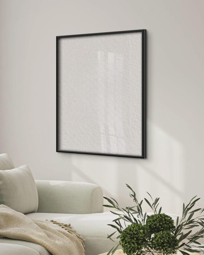 May include: A square wall-mounted picture frame with a thin black border and a blank white interior surface. The frame is positioned on a plain wall above a light-colored sofa, with a small green plant visible in the foreground of the scene.