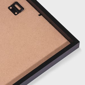 May include: The back of a black rectangular picture frame featuring a brown fiberboard backing. A black metal sawtooth hanger is attached to the upper left corner of the frame, providing a secure point for wall mounting the display item.