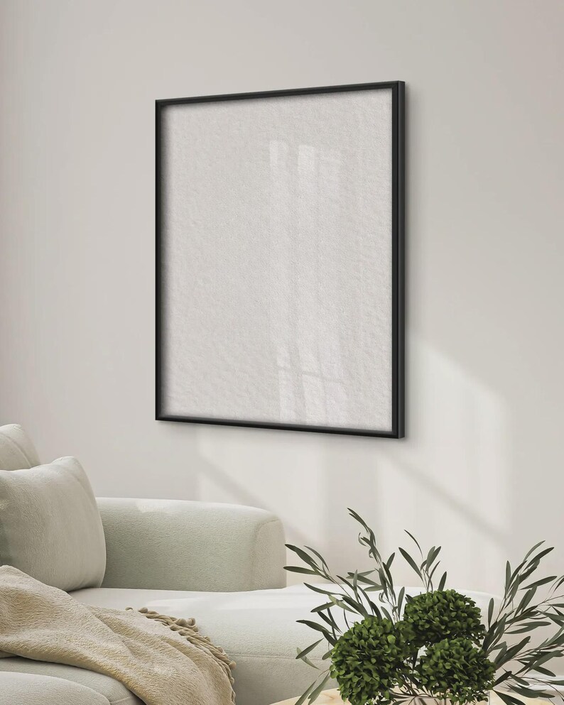 May include: A square wall-mounted picture frame with a thin black border and a blank white interior surface. The frame is positioned on a plain wall above a light-colored sofa, with a small green plant visible in the foreground of the scene.