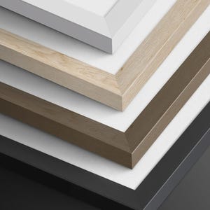 May include: A stack of four rectangular picture frames with mitered corners. The frames are arranged in a descending order, featuring a smooth white finish, a light natural wood grain, a medium brown wood grain, and a solid matte black finish.