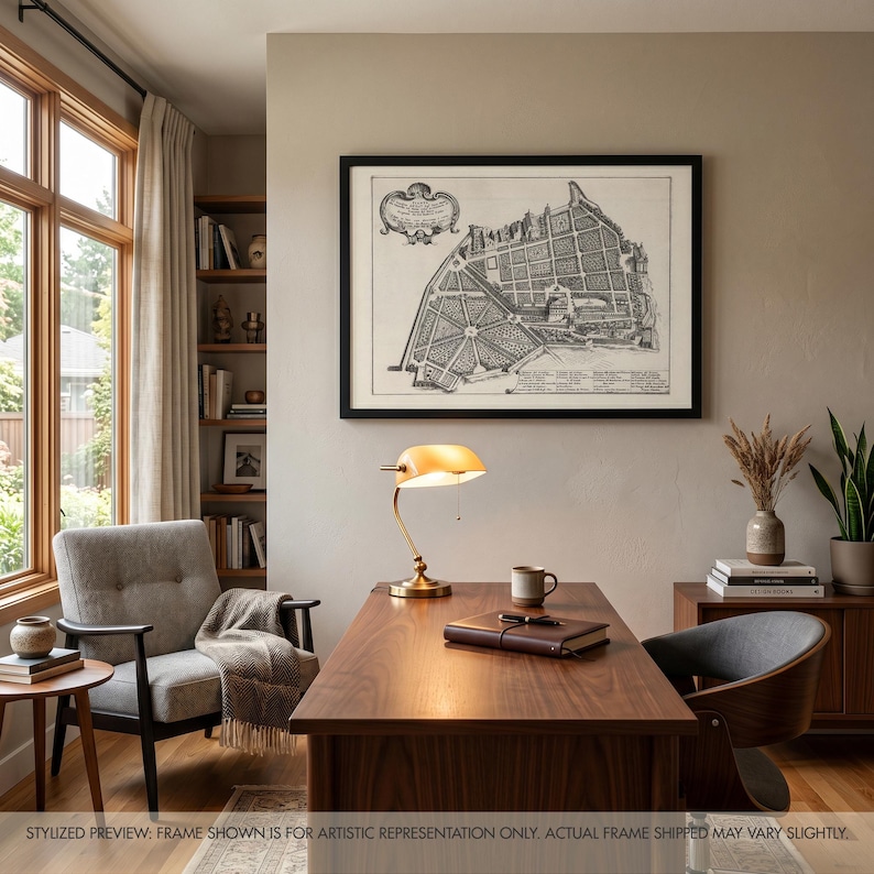 May include: A black framed wall art print featuring a detailed historical map of a garden layout with intricate line work and text, displayed above a wooden desk with a brass desk lamp, a brown leather notebook, and a ceramic mug.