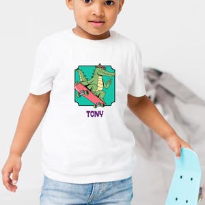 Könnte beinhalten: Ein Kind trägt ein weißes T-Shirt mit einem Grafik eines Alligators auf einem Skateboard und dem Namen "TONY". Das Kind hält ein hellblaues Skateboard mit rosa Rollen. Es trägt eine blaue Jeans und braune Schuhe.