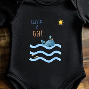 Può includere: Body per neonati nero con un'illustrazione di balena. Il design include il testo "CALVIN IS ONE", una balena blu che spruzza acqua, linee blu ondulate, un sole giallo e piccoli pesci.