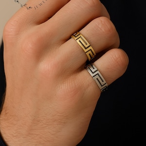 Könnte beinhalten: Zwei Ringe mit griechischem Schlüsselmuster werden auf einer Hand gezeigt. Ein Ring ist goldfarben, der andere silberfarben. Die Ringe sind schlichte Bänder mit einem sich wiederholenden geometrischen Muster. Die Ringe sind Teil der Schmuckkollektion von Bold Bling Co.