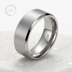 Può includere: Un anello in titanio color argento con finitura spazzolata. L'anello ha una parte superiore piatta e bordi smussati. La parola "TITANIUM" è incisa all'interno. L'anello è esposto su una superficie bianca.