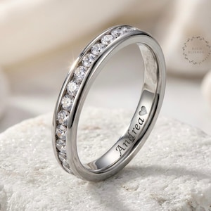 May include: A silver-toned metal band ring featuring a channel setting with a row of clear, round-cut stones. The outer surface of the ring is engraved with the name Andrea followed by a small heart symbol in a script-style font.
