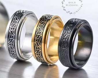 May include: Three stainless steel spinner rings in silver, gold, and black. Each ring features a Celtic knot design around the band. The rings are displayed on a reflective surface. The brand name "BOLD BLINGCO. JEWELRY" is in a circular design.