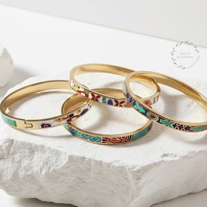 Può includere: Quattro braccialetti dorati con disegni smaltati colorati. I braccialetti presentano motivi in rosso, blu, bianco e turchese. Un braccialetto ha una chiusura. I gioielli sono esposti su una superficie bianca strutturata.