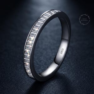 May include: A dark-toned metal band ring featuring a channel setting of clear, rectangular baguette-cut stones along the outer surface. The interior of the band is clearly engraved with the text S925, indicating sterling silver composition.