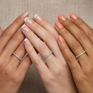 May include: Three thin eternity bands featuring small round-cut clear stones set in metal. One ring is silver-toned, one is rose gold-toned, and one is yellow gold-toned. Each band is worn on a finger, showcasing the stones set along the entire circumference.