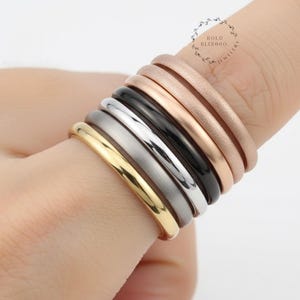 Könnte beinhalten: Eine Auswahl an stapelbaren Ringen in verschiedenen Farben, darunter Gold, Silber, Schwarz und Roségold. Die Ringe werden an einem Finger präsentiert und zeigen ihre unterschiedlichen Oberflächen und Texturen. Der Schmuck ist schlicht und elegant.