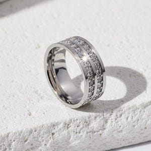 Puede incluir: Un anillo plateado con dos filas de pequeñas piedras preciosas brillantes. El anillo está sobre una superficie blanca y texturizada, con una sombra proyectada hacia la derecha. El anillo parece ser de metal brillante.