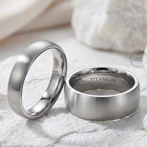 May include: Two silver titanium rings. One ring is upright, displaying the word "TITANIUM" engraved on the inside. The other ring is on its side, with the word "TITANIUM" engraved on the inside. The rings are on a white, textured surface.