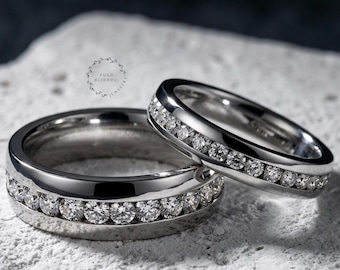 May include: Two silver rings with a row of sparkling clear stones set into the band. One ring is wider than the other. The rings are on a textured white surface.