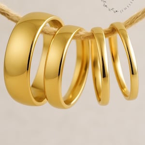 May include: Four gold-colored rings of varying widths are suspended from a natural-colored rope. The rings have a polished, reflective surface. The rings are labeled with the text "BOLD BLING&CO. JEWELRY."