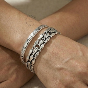 Op de afbeelding: Twee zilveren armbanden met vismotieven. Eén armband is een eenvoudige band met gegraveerde vissen, de andere heeft meerdere vissen in rijen. De armbanden worden om een pols gedragen, waarbij de ingewikkelde details van de sieraden worden getoond.