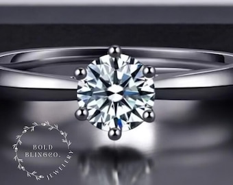 May include: A close-up of a silver ring with a large, clear, round-cut diamond. The diamond is set with six prongs. The ring band is smooth and polished. The ring is set against a dark background. The brand name "BOLD BLING CO. JEWELLERY" is in the lower left corner.