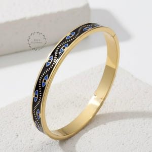May include: Gold-coloured bangle bracelet with a black enamel band featuring blue eye designs. The bracelet has a hinged clasp and a polished finish. The jewellery is displayed on a white surface.